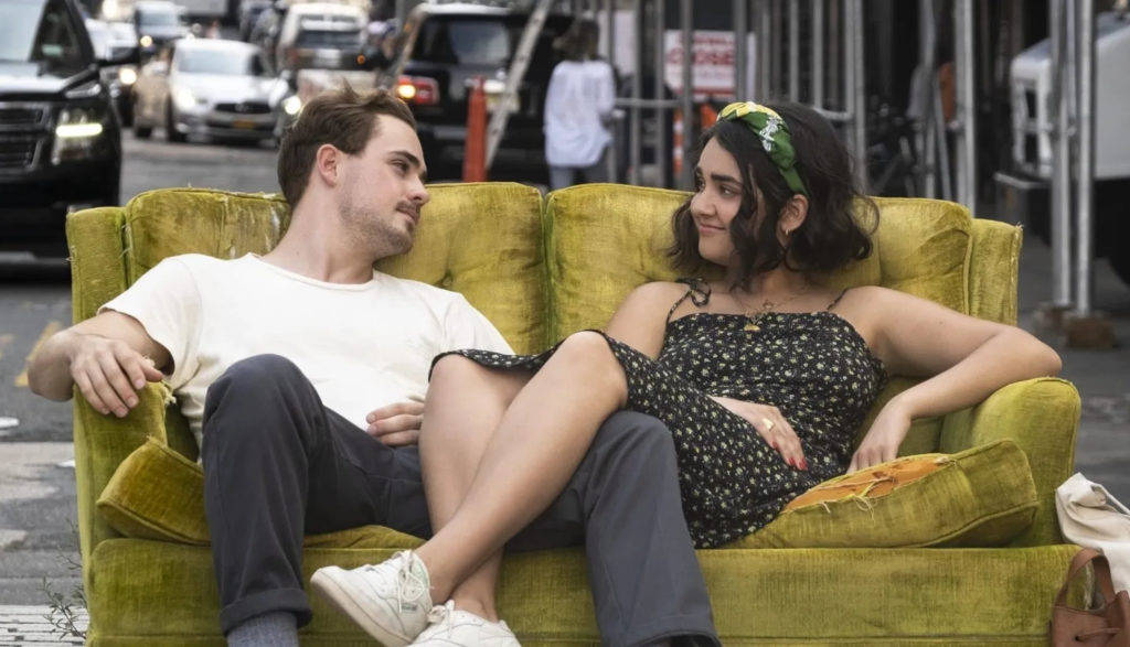 A young man and woman sit on a couch together in the middle of a busy city street.