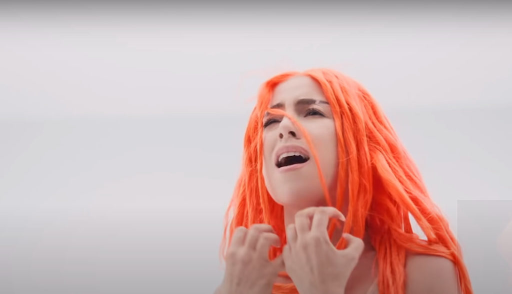 Orange-haired singer Ava Max clenches her hands as she sings in apparent emotional agony.