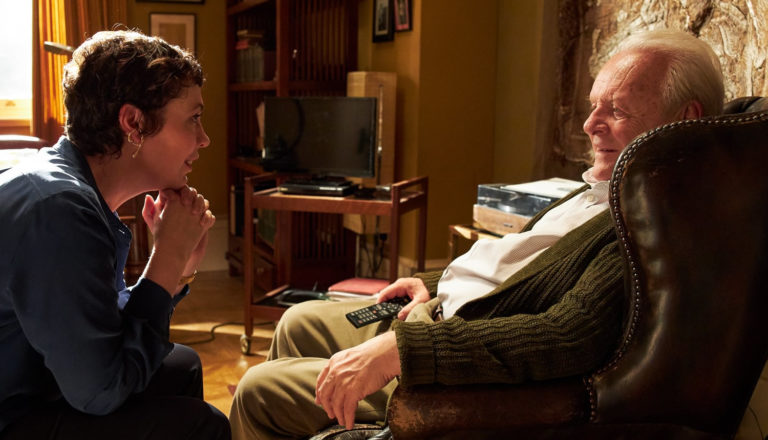 An elderly man sits in a chair and talks to his middle-aged daughter.
