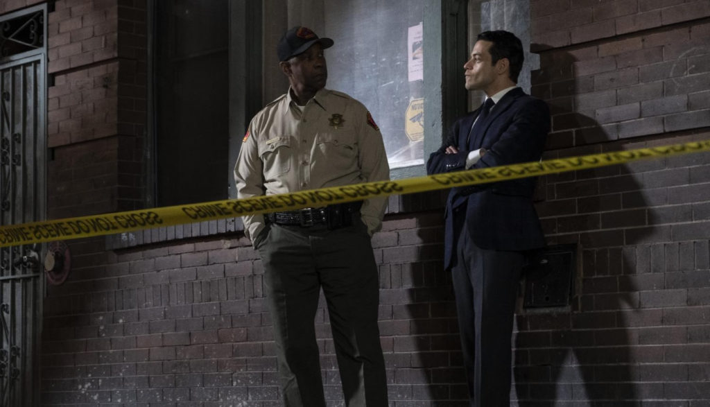 A police officer and a detective stand near a building and a crime scene.