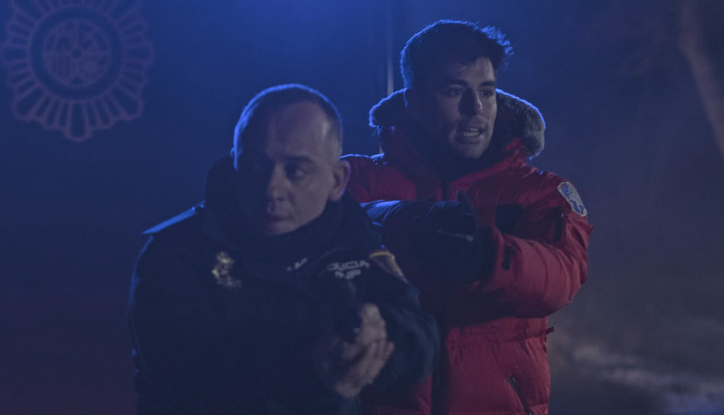 Two men with guns drawn walk through a dark, cold outside area.