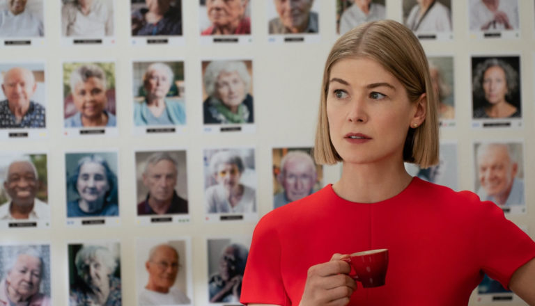 A woman stands with a cup of tea in front of portrait photos of elderly people behind her.