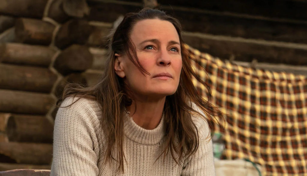 A woman (played by actress Robin Wright) sits in front of a log cabin with a thoughtful expression on her face.