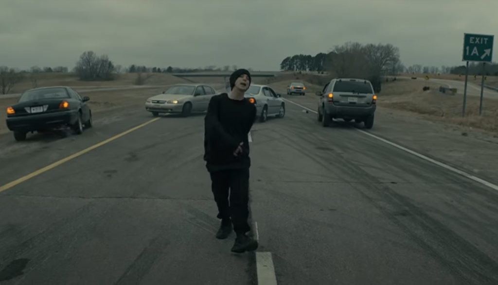 The Christian rapper NF walks down the middle of an interstate highway as cars swerve to avoid him.