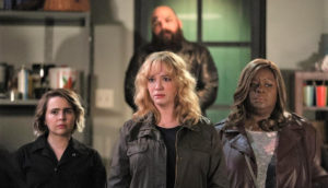 Three middle aged women have concerned looks on their faces as an ominous man lurks behind them.