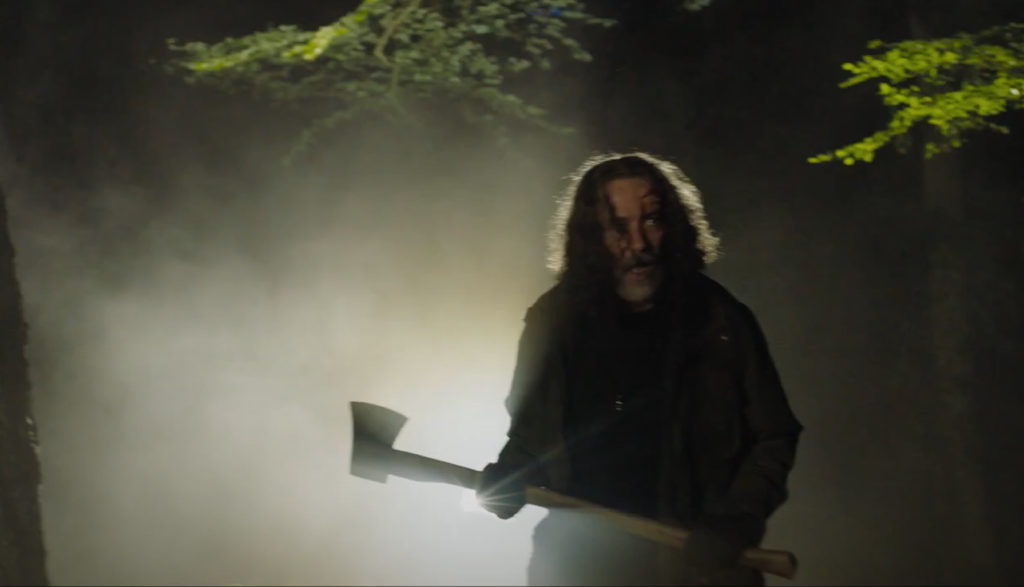 A creepy man with an ax walks through a poorly lit forest.