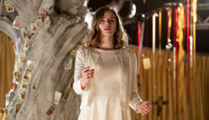 A woman in a white dress stands in front of a tree with many pictures wedged into it.