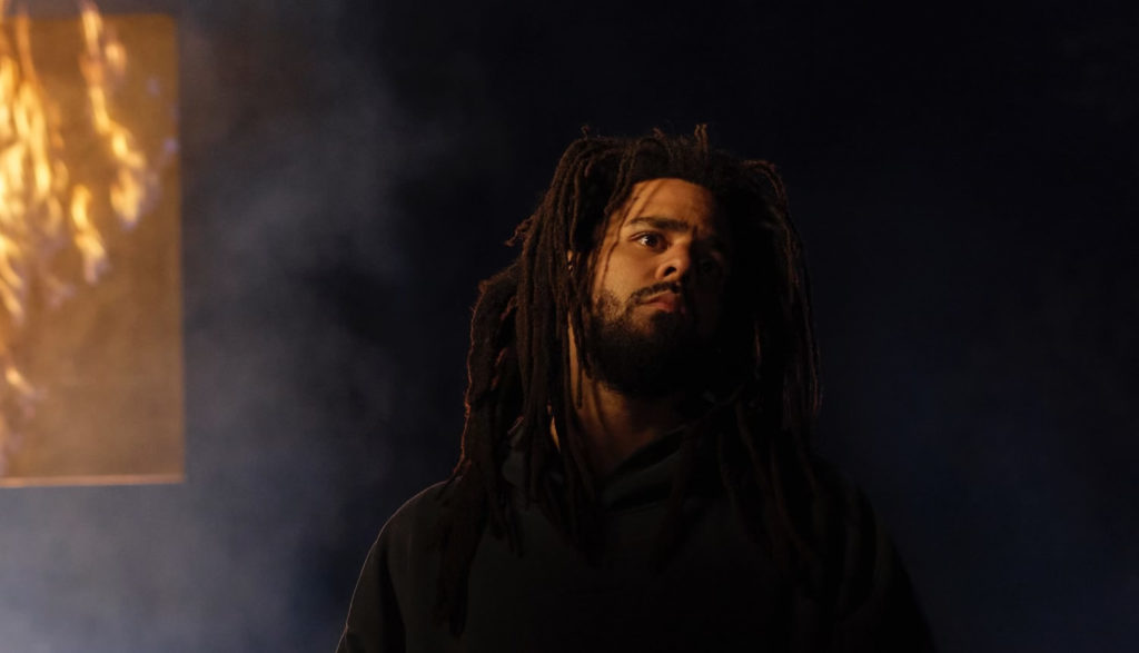 Picture of rapper J. Cole in a dark space superimposed against another image of fire.