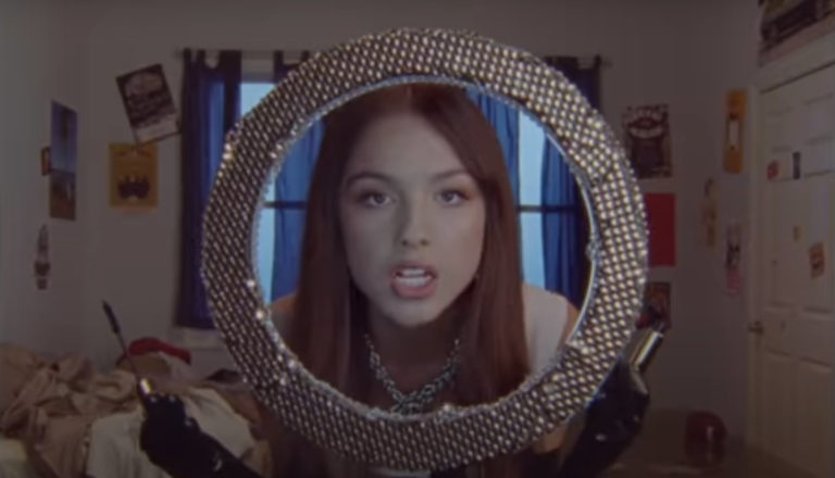 Picture of singer Olivia Rodrigo singing into a mirror, where we see the reflected image of her angry face.