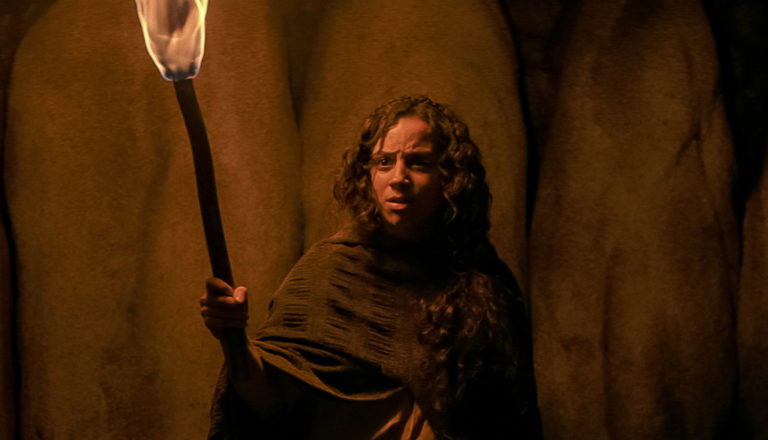 A person in a dark space holds a torch aloft and looks quite concerned.