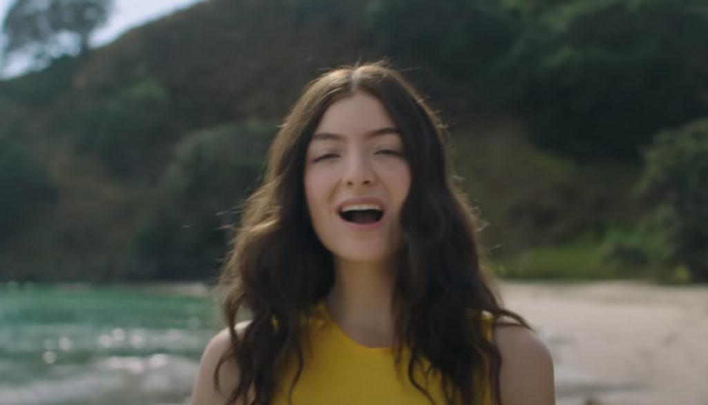 Pop artist Lorde sings on a beach in a yellow dress.