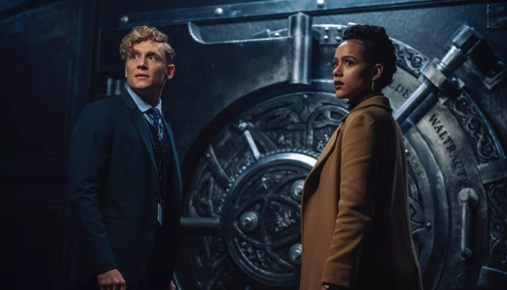 A young man and young woman stand in front of a huge safe that they're trying to crack.