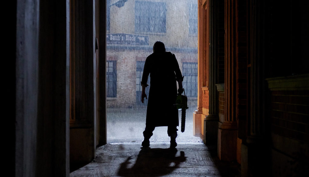 We see the silhouette of a man holding a chainsaw in a dark, rainy alley.