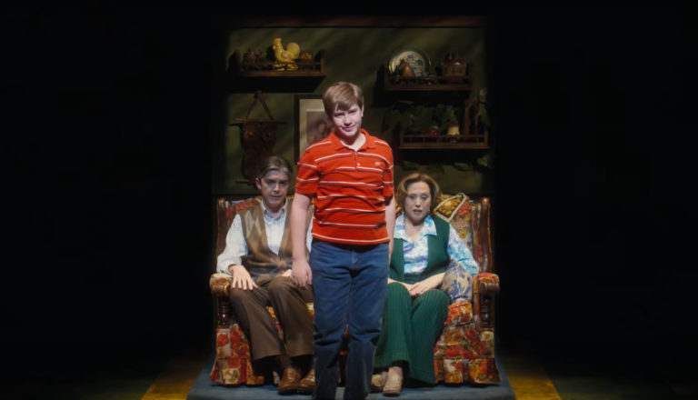 A smiling boy stands in front of his obviously weary parents, who are sitting down.