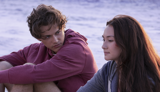 A young couple sit talking on a beach, looking pensive.