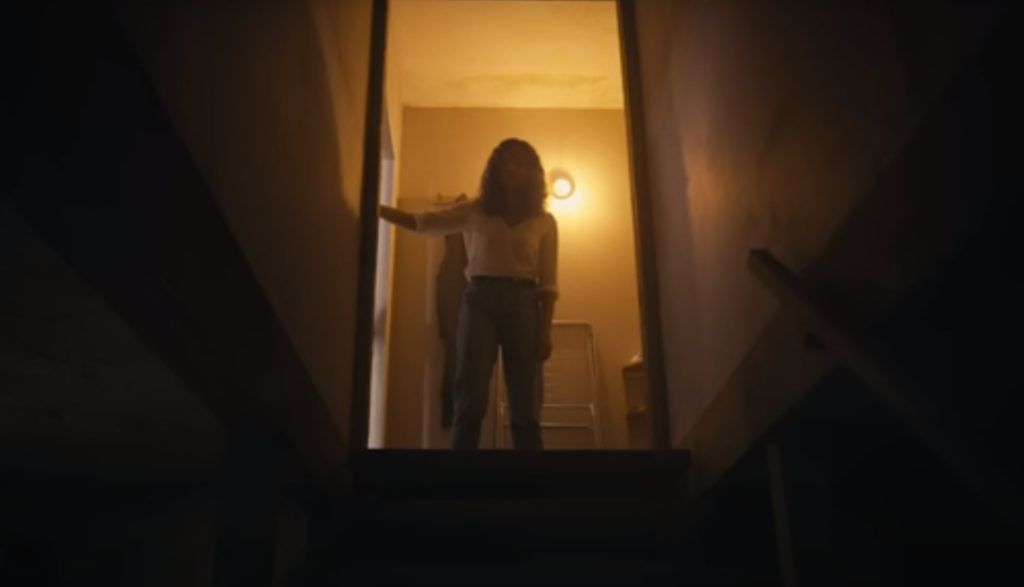 A woman stands atop a staircase and looks down into a freakishly dark basement.
