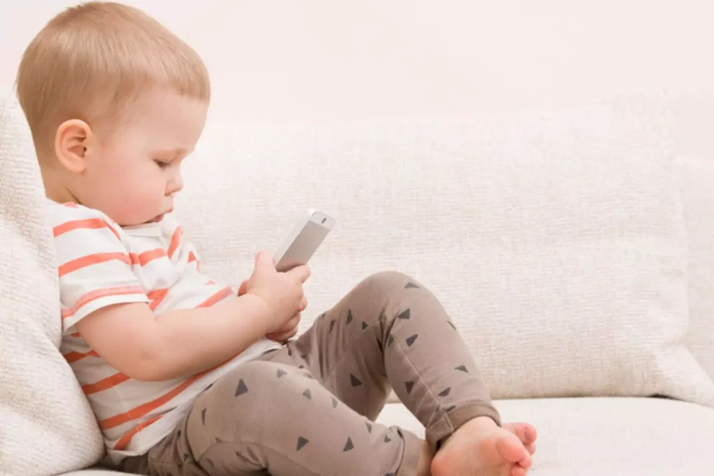 Addicted to screens- a toddler addicted to screens is sitting on the couch holding a smartphone|Can babies get addicted to screens?|Addicted to screens