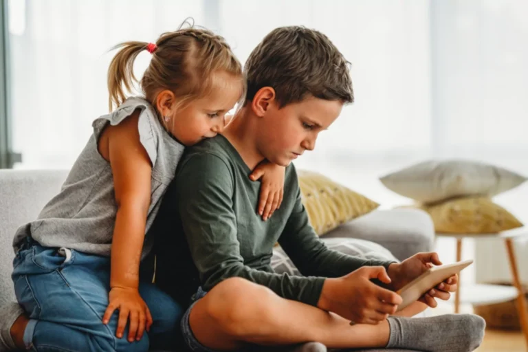 A young boy watching a tablet closely while a younger girl leans over his shoulder