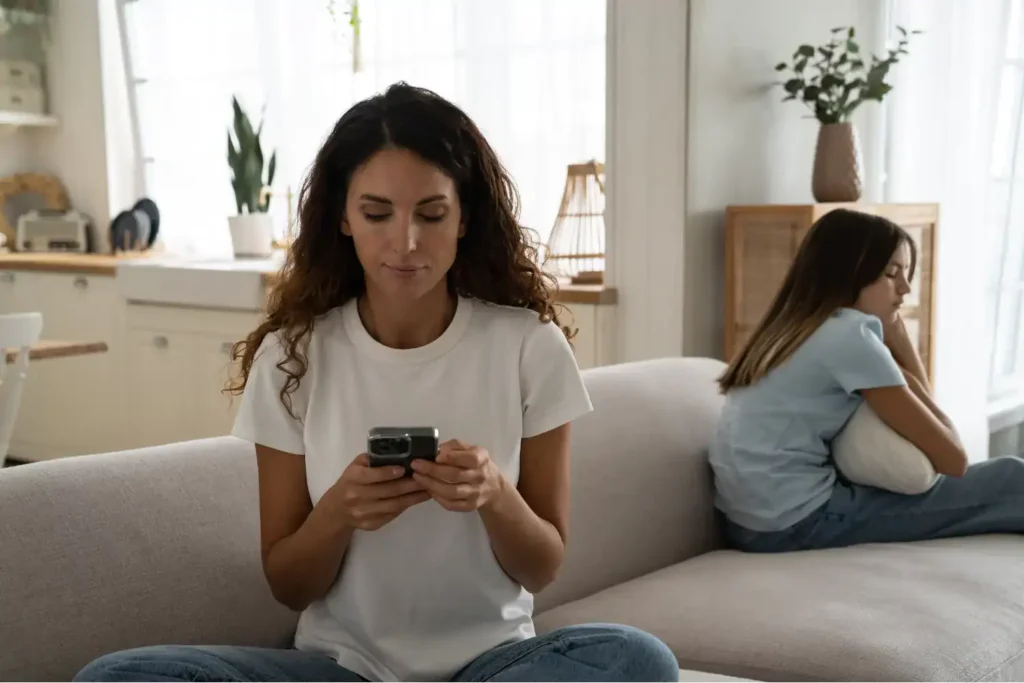 Mother focused on her phone while daughter sits upset beside her